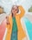 A fashionable woman wearing a bright yellow coat over a green top and blue jeans stands confidently with one hand in her hair. She wears stylish round sunglasses and large colorful bracelets, set against the backdrop of a playground slide with vibrant red and blue colors.