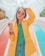 A fashionable woman wearing a bright yellow coat over a green top and blue jeans stands confidently with one hand in her hair. She wears stylish round sunglasses and large colorful bracelets, set against the backdrop of a playground slide with vibrant red and blue colors.