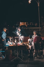 A night market scene featuring a group of people interacting around a lit-up vendor's table, displaying various items. The area is illuminated by artificial lighting, creating a contrast with the dark surroundings. An American flag is seen hanging above the table, and some individuals are browsing or conversing.