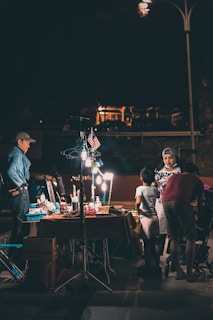 A night market scene featuring a group of people interacting around a lit-up vendor's table, displaying various items. The area is illuminated by artificial lighting, creating a contrast with the dark surroundings. An American flag is seen hanging above the table, and some individuals are browsing or conversing.