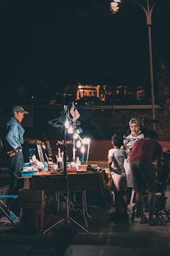 A night market scene featuring a group of people interacting around a lit-up vendor's table, displaying various items. The area is illuminated by artificial lighting, creating a contrast with the dark surroundings. An American flag is seen hanging above the table, and some individuals are browsing or conversing.