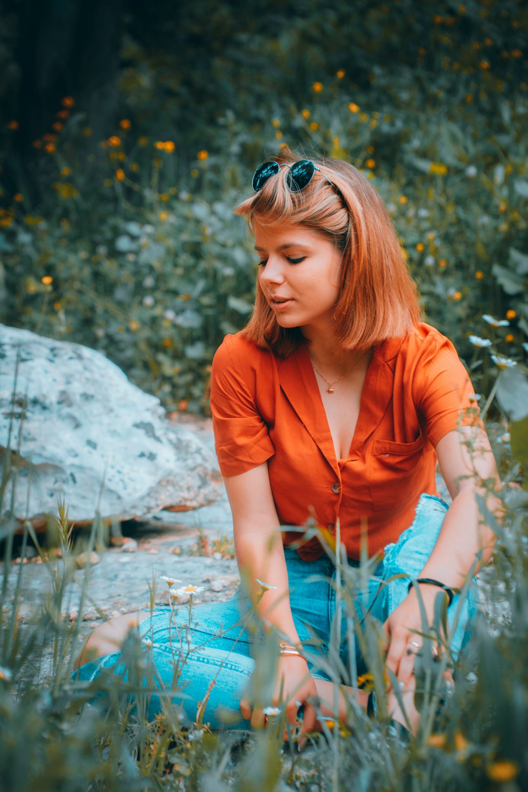 woman in orange button up shirt wearing blue sunglasses sitting on rock in river during daytime