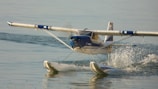 A seaplane is skimming across the water's surface, creating a trail of splashes behind it. The aircraft's wings are extended and pontoons are visible, designed to allow for water landings and takeoffs. The sun reflects off the water and the plane's metallic surfaces, highlighting the graceful movement.