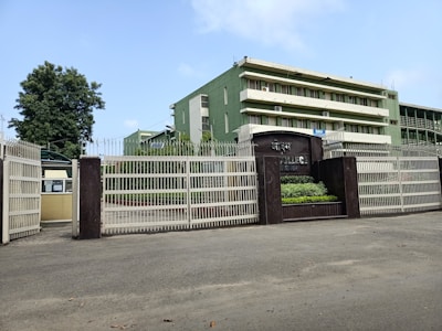 A building with multiple stories painted in green, surrounded by a large metal fence. A tree is visible to the left, and there is a sign indicating the name of the institution. The sky is blue with a few clouds.