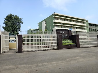 A building with multiple stories painted in green, surrounded by a large metal fence. A tree is visible to the left, and there is a sign indicating the name of the institution. The sky is blue with a few clouds.