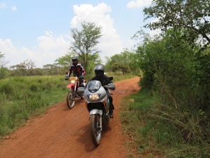 Two motorcyclists wearing helmets and protective gear ride on a dirt path surrounded by greenery and trees under a partly cloudy blue sky.