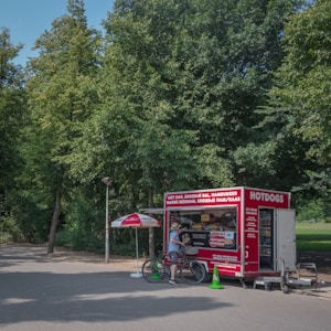 A food stall with a red exterior offering items like hot dogs and hamburgers, located in a park setting. A person stands next to the stall with a bicycle, conversing with the vendor. An umbrella provides shade to the customer area, surrounded by lush green trees.