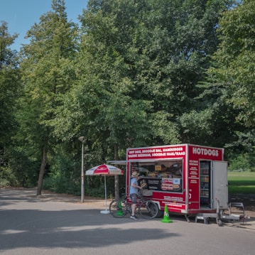A food stall with a red exterior offering items like hot dogs and hamburgers, located in a park setting. A person stands next to the stall with a bicycle, conversing with the vendor. An umbrella provides shade to the customer area, surrounded by lush green trees.