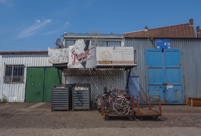 A variety of metal buildings with different colored doors, including green and blue, and a collection of old bicycles stacked together. There is a sign displaying the words 'Café Joost' and another about recycling services. The area seems industrial and slightly worn, with a paved ground and blue sky above.