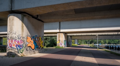 Urban environment with large concrete bridge supports covered in colorful graffiti art. The bridge stretches overhead, casting shadows on the pathway below, while greenery and trees are visible in the background. This area appears to be a pedestrian or cycling path adjacent to a body of water.