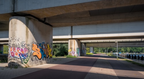Urban environment with large concrete bridge supports covered in colorful graffiti art. The bridge stretches overhead, casting shadows on the pathway below, while greenery and trees are visible in the background. This area appears to be a pedestrian or cycling path adjacent to a body of water.