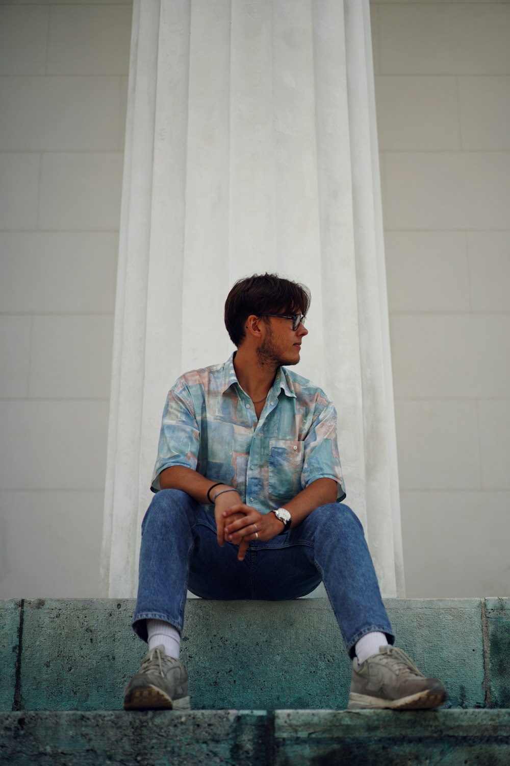 man in blue and white button up shirt and blue denim jeans sitting on white concrete
