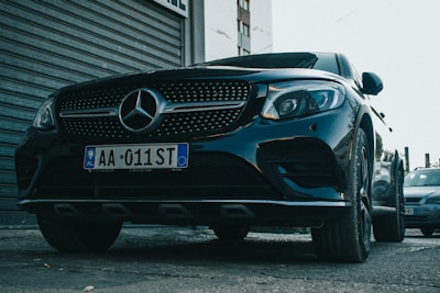A sleek black Mercedes-Benz car is parked on a street, with the front grille prominently displaying the iconic Mercedes star emblem. The vehicle's polished surface reflects the surrounding environment, and the license plate reads AA 011 ST. The background includes a corrugated metal door and part of a building.