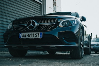 A sleek black Mercedes-Benz car is parked on a street, with the front grille prominently displaying the iconic Mercedes star emblem. The vehicle's polished surface reflects the surrounding environment, and the license plate reads AA 011 ST. The background includes a corrugated metal door and part of a building.