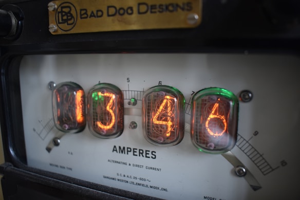 A close-up view of a vintage device displaying numerical readings on glowing tubes, marked with the label 'Bad Dog Designs.' The readout shows the numbers 1346 and the word 'Amperes.' The device appears to measure alternating and direct current.
