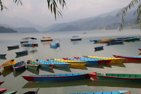 A serene lake scene featuring numerous colorful boats floating on calm water. The boats, painted in vibrant hues such as blue, yellow, red, and green, are arranged across the lake. The background displays misty mountains under a cloudy sky, creating a sense of depth and tranquility. Overhanging leaves frame the top edge of the image, adding a touch of natural detail.