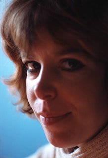 A close-up portrait of a person with short, wavy brown hair and a light complexion. The background is a soft, muted blue, which contrasts with the warm tones of the subject's face and hair. The person has an introspective expression with softly lit features, and they are wearing a cream-colored knit garment.