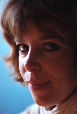 A close-up portrait of a person with short, wavy brown hair and a light complexion. The background is a soft, muted blue, which contrasts with the warm tones of the subject's face and hair. The person has an introspective expression with softly lit features, and they are wearing a cream-colored knit garment.