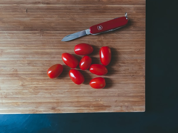 A wooden cutting board with a red-handled Swiss Army knife and nine small red tomatoes arranged on it.