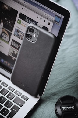 A smartphone with a black case is leaning against a laptop, which displays a YouTube page. The laptop keyboard is visible, and a camera lens cap labeled 'YONGNUO DIGITAL' is nearby.