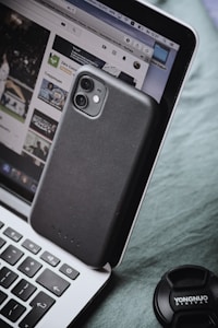 A smartphone with a black case is leaning against a laptop, which displays a YouTube page. The laptop keyboard is visible, and a camera lens cap labeled 'YONGNUO DIGITAL' is nearby.