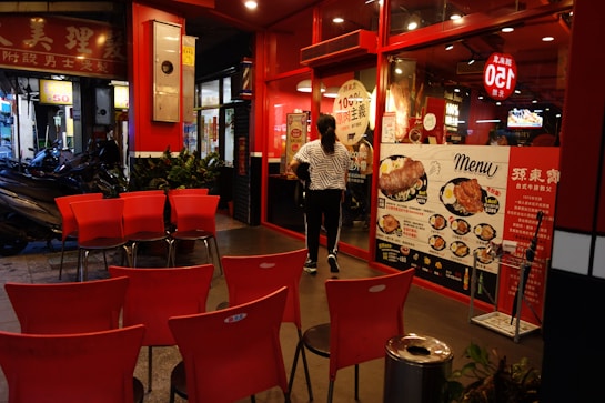 An urban dining area with red chairs arranged outside a restaurant. The facade is decorated with vibrant red and black, featuring large menu displays with images of various dishes. A person in a striped shirt is walking towards the entrance of the restaurant. Motorbikes are parked on the side of the walkway.