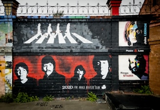 A mural painted on a brick wall depicts iconic images associated with The Beatles. The top section shows silhouettes of four people in a formation walking on a crosswalk, reminiscent of the Abbey Road album cover. Below, four stylized portraits of The Beatles members are painted in black on a red background. To the right, there are colorful pop-art style portraits of two individuals with accompanying text reading 'John Lennon' and 'Ringo Starr'.