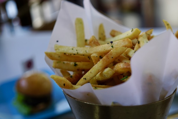 A serving of golden-brown French fries is placed upright in a metal cone lined with white paper. The fries are seasoned with herbs and black pepper. In the background, there is a blurred view of a hamburger on a blue plate.