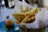 A serving of golden-brown French fries is placed upright in a metal cone lined with white paper. The fries are seasoned with herbs and black pepper. In the background, there is a blurred view of a hamburger on a blue plate.