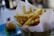 A serving of golden-brown French fries is placed upright in a metal cone lined with white paper. The fries are seasoned with herbs and black pepper. In the background, there is a blurred view of a hamburger on a blue plate.