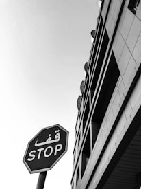 A black and white photograph featuring a stop sign with both Arabic and English text. The sign is positioned on the left side of the image, set against the backdrop of a modern building with geometric architectural features. The sky is clear, adding emphasis to the stark contrast and lines.