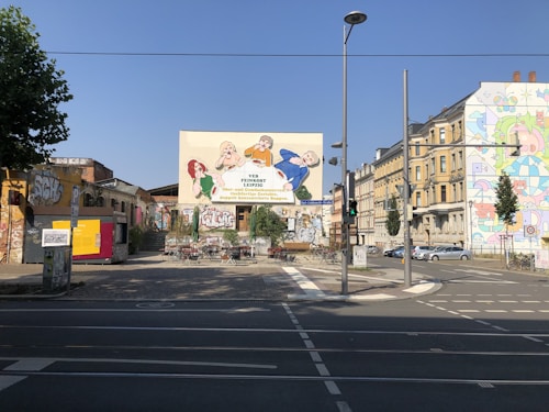 An urban street scene featuring buildings with colorful murals and graffiti. In the center, there is a prominent wall with a large mural depicting cartoon characters. The area has outdoor seating, possibly for a cafe, with tables and chairs scattered around. Street signs and a traffic light are visible, along with parked cars on the right side.