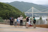A group of people stand facing a scenic view with lush green hills and a large cable-stayed bridge in the background. They appear to be tourists, some taking photos and others simply observing the landscape. The scene suggests a peaceful and leisurely outing.