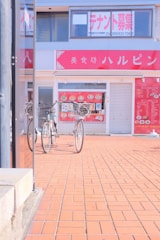 Two bicycles are parked in front of a storefront with large glass windows and red signage displaying Japanese characters. The ground is paved with red tiles, and a menu is visible on the storefront window.