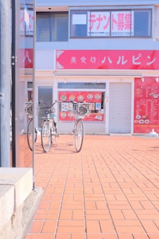 Two bicycles are parked in front of a storefront with large glass windows and red signage displaying Japanese characters. The ground is paved with red tiles, and a menu is visible on the storefront window.