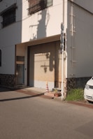 A closed garage door in a residential or small commercial building with a yellow and orange traffic cone placed in front of it. The shadows of utility poles are cast on the wall in the late afternoon light. There are two potted plants next to the door and part of a white vehicle is visible on the right. The exterior wall displays a mix of smooth and textured finishes.