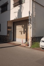 A closed garage door in a residential or small commercial building with a yellow and orange traffic cone placed in front of it. The shadows of utility poles are cast on the wall in the late afternoon light. There are two potted plants next to the door and part of a white vehicle is visible on the right. The exterior wall displays a mix of smooth and textured finishes.