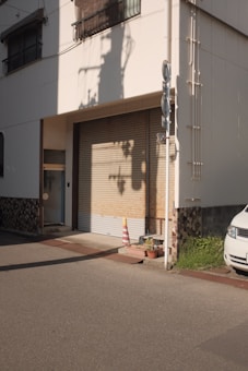 A closed garage door in a residential or small commercial building with a yellow and orange traffic cone placed in front of it. The shadows of utility poles are cast on the wall in the late afternoon light. There are two potted plants next to the door and part of a white vehicle is visible on the right. The exterior wall displays a mix of smooth and textured finishes.
