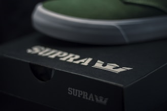 A close-up of a black shoe box with the brand name 'SUPRA' and a crown logo in white. The upper edge of a green shoe with a white sole is visible, resting on the box.