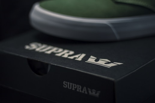 A close-up of a black shoe box with the brand name 'SUPRA' and a crown logo in white. The upper edge of a green shoe with a white sole is visible, resting on the box.