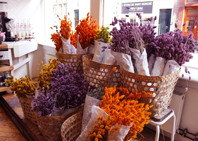 A collection of vibrant dried flowers arranged in wicker baskets. The flowers are predominantly in shades of orange, yellow, and purple, wrapped in white paper. The baskets are placed in a cozy, well-lit room with wooden floors and a counter with various items in the background.
