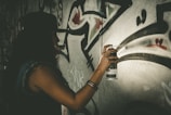 A person wearing a hat is creating graffiti art on a wall using a spray paint can. The wall features abstract shapes and bold, sweeping lines.