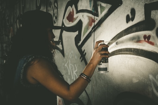 A person wearing a hat is creating graffiti art on a wall using a spray paint can. The wall features abstract shapes and bold, sweeping lines.