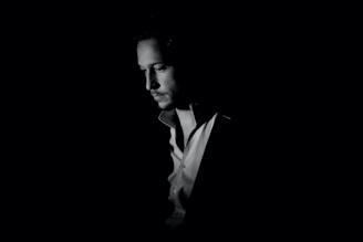A person is shown in a black and white portrait with dramatic lighting, highlighting the facial features and casting deep shadows. The focus is on the introspective expression.