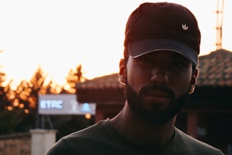 A person wearing a dark cap stands in front of a sunset background with trees and a blurred sign that reads 'ETAC'. The setting sun creates a silhouette effect, highlighting the contours of the person's face.