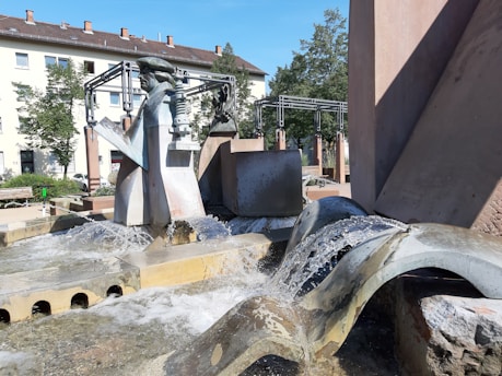A modern, urban water fountain with various geometric concrete structures and metallic sculptures from which water flows. There are grey and brown cubic shapes alongside curved water paths. In the background, there are buildings, green trees, and benches. The sky is clear, indicating a sunny day.