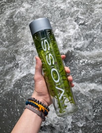 A hand is holding a clear bottle filled with water and green plant stems inside. The label on the bottle is visible, and it features a gray cap. In the background, water is flowing energetically, creating a dynamic and refreshing scene. The person wearing colorful bracelets on their wrist adds a touch of vibrancy.