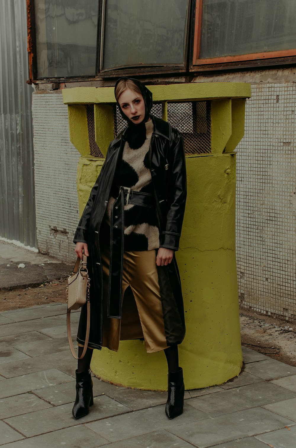 woman in black coat and gray pants standing beside yellow concrete wall