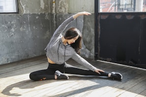 A person is seated on a wooden floor in a spacious room doing a side stretch. They are wearing a gray hoodie and black leggings with athletic shoes. The walls have a rustic, worn appearance with peeling paint, and there are large windows allowing natural light to illuminate the space.