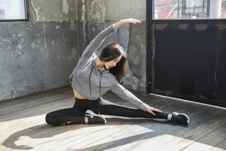 A person is seated on a wooden floor in a spacious room doing a side stretch. They are wearing a gray hoodie and black leggings with athletic shoes. The walls have a rustic, worn appearance with peeling paint, and there are large windows allowing natural light to illuminate the space.
